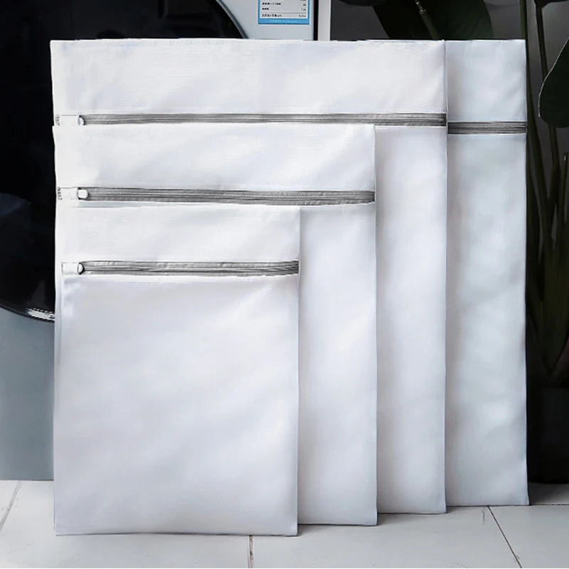 Set of four white mesh laundry bags with gray zippers in assorted sizes in front of a washing machine