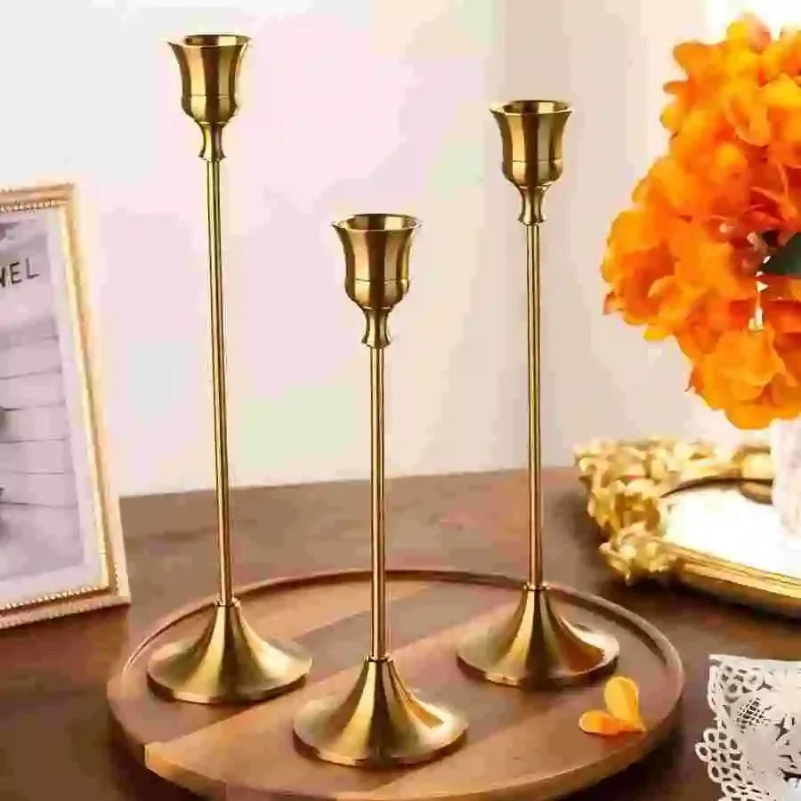 Set of three gold metal candlestick holders displayed on a wooden tray with bright orange flowers and a framed sign in the background.