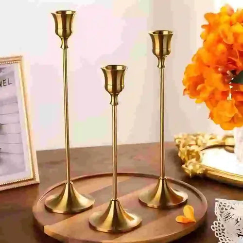 Set of three gold metal candlestick holders displayed on a wooden tray with bright orange flowers and a framed sign in the background.