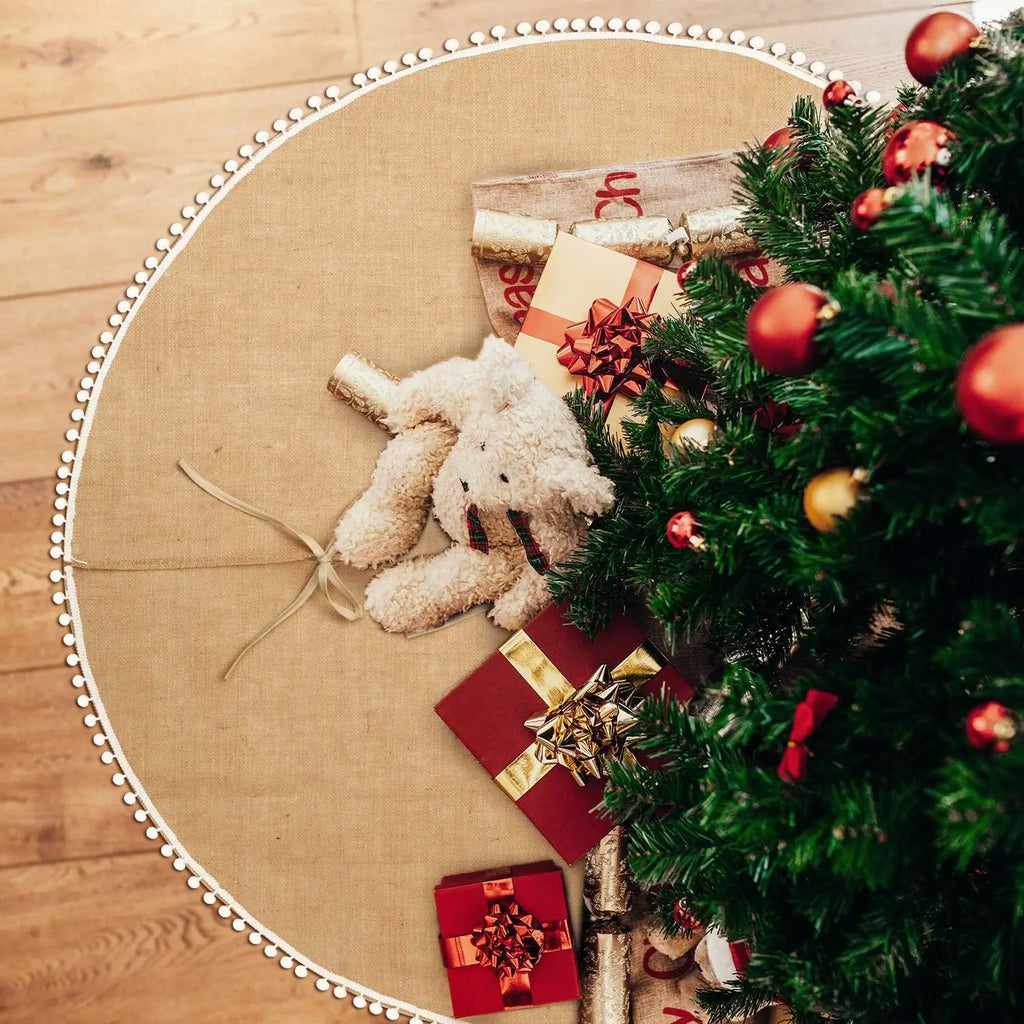 Round burlap tree skirt with pompom trim holding presents and plush toy under tree