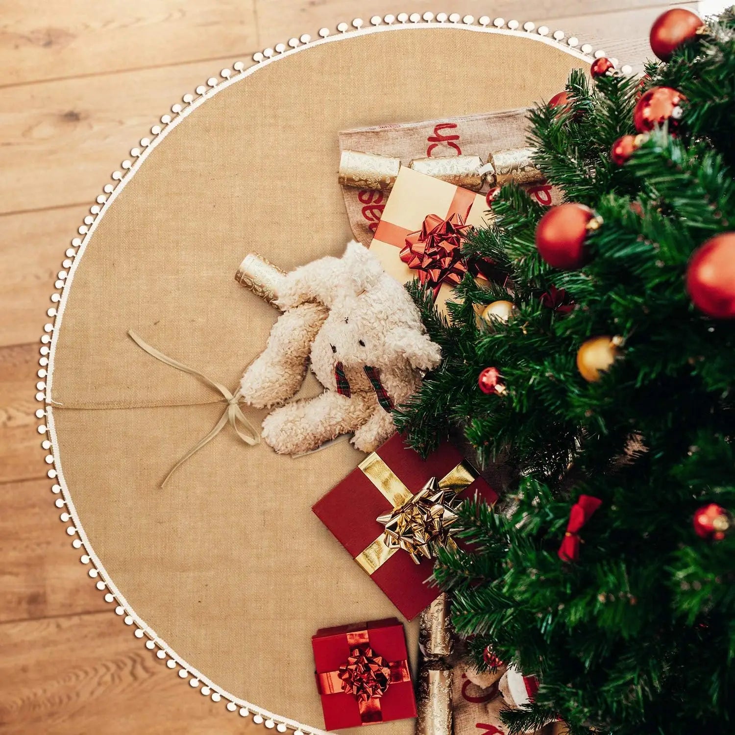 Round burlap tree skirt with pompom trim holding presents and plush toy under tree