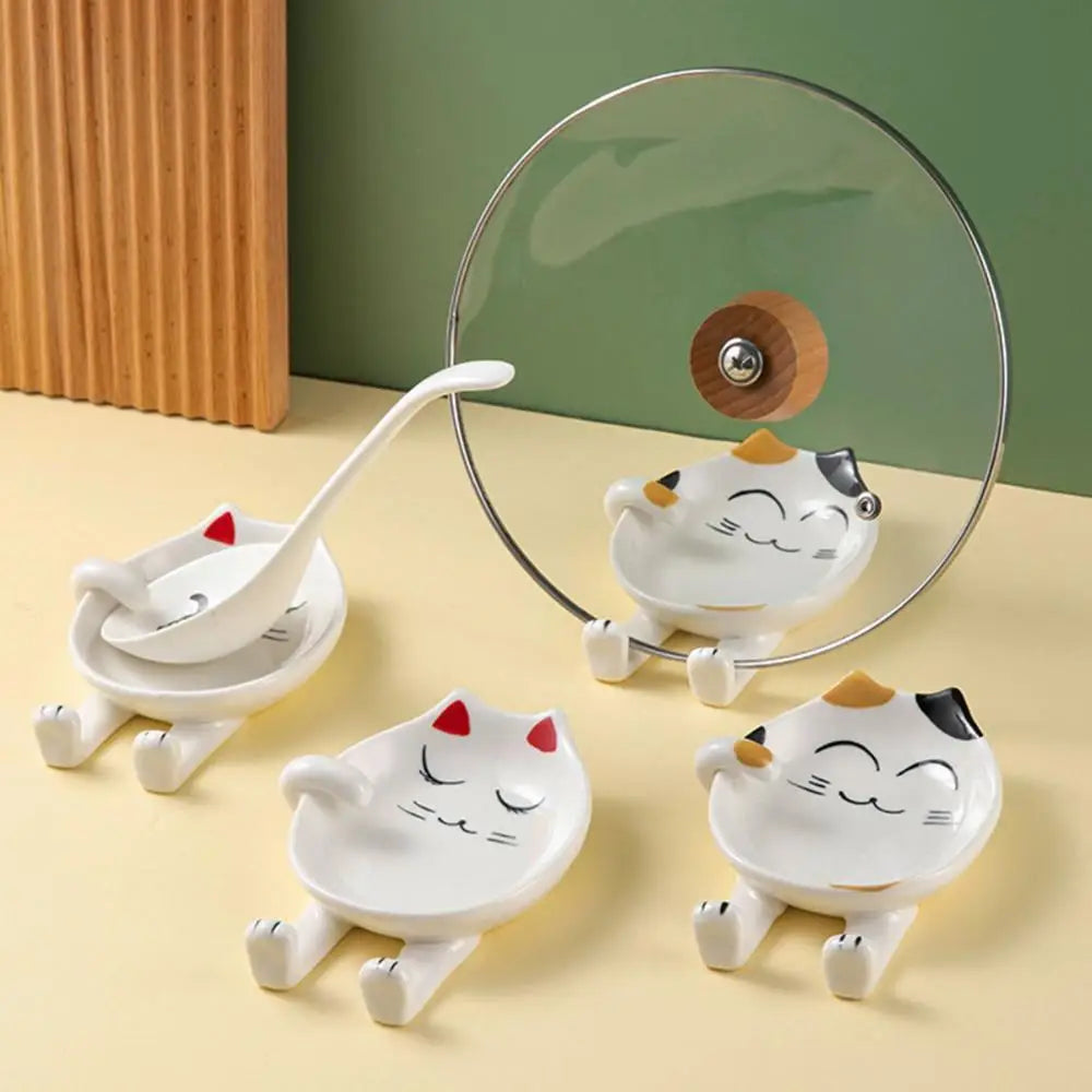 Two ceramic cat-shaped spoon rests in yellow and red styles, shown holding a ladle and pot lid on a kitchen counter