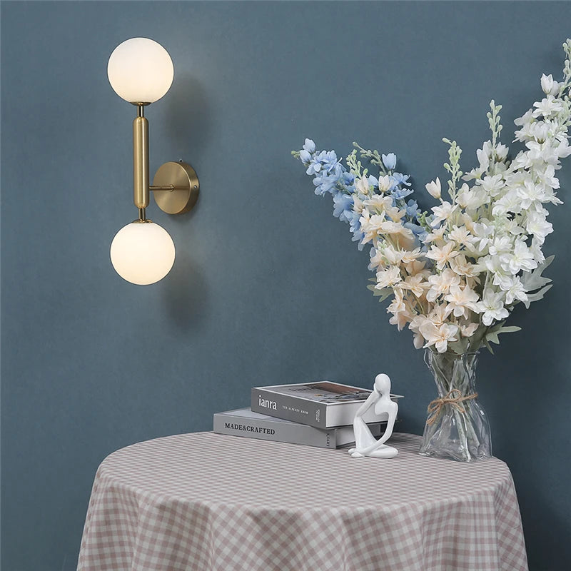 Vertical two globe wall lamp in gold finish on blue wall beside checkered tablecloth and flowers