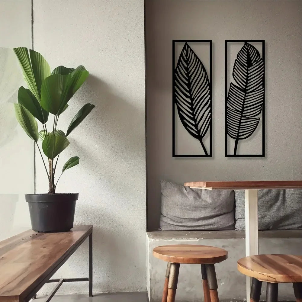 Set of two black metal leaf wall art panels hung on a light wall above a bench and table