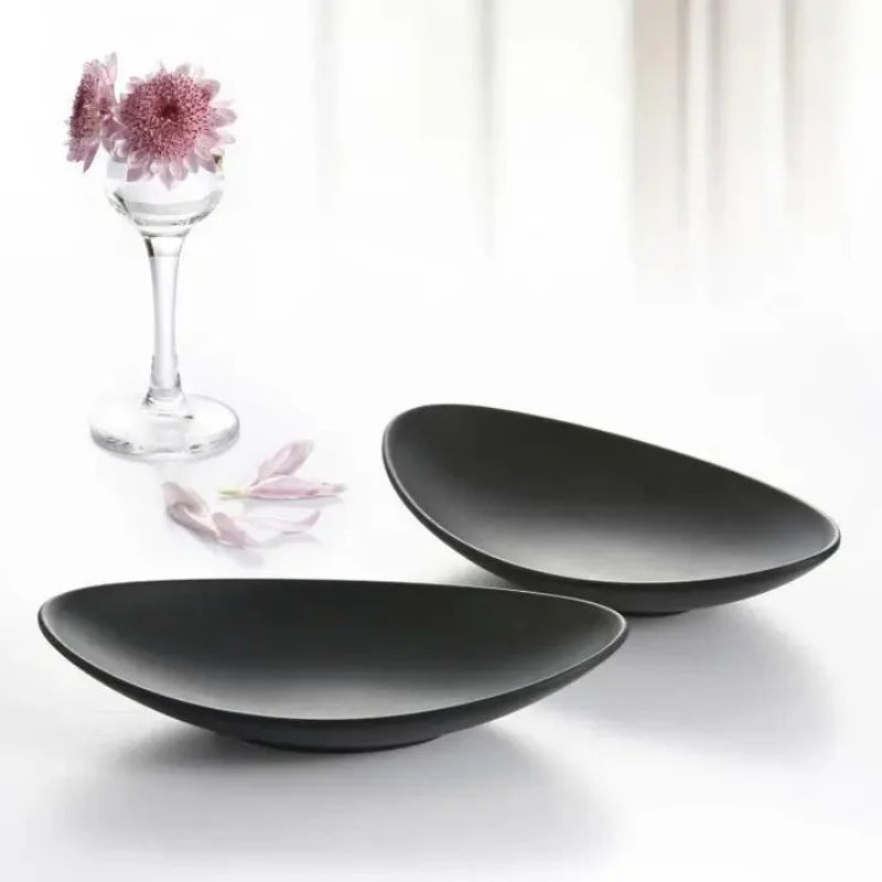 pair of matte black oval ceramic trays with a glass vase and pink flower petals against a light background
