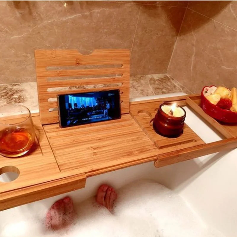 Bamboo bathtub tray in use holding a drink, candle, phone, fruit, and with feet visible in bath water.