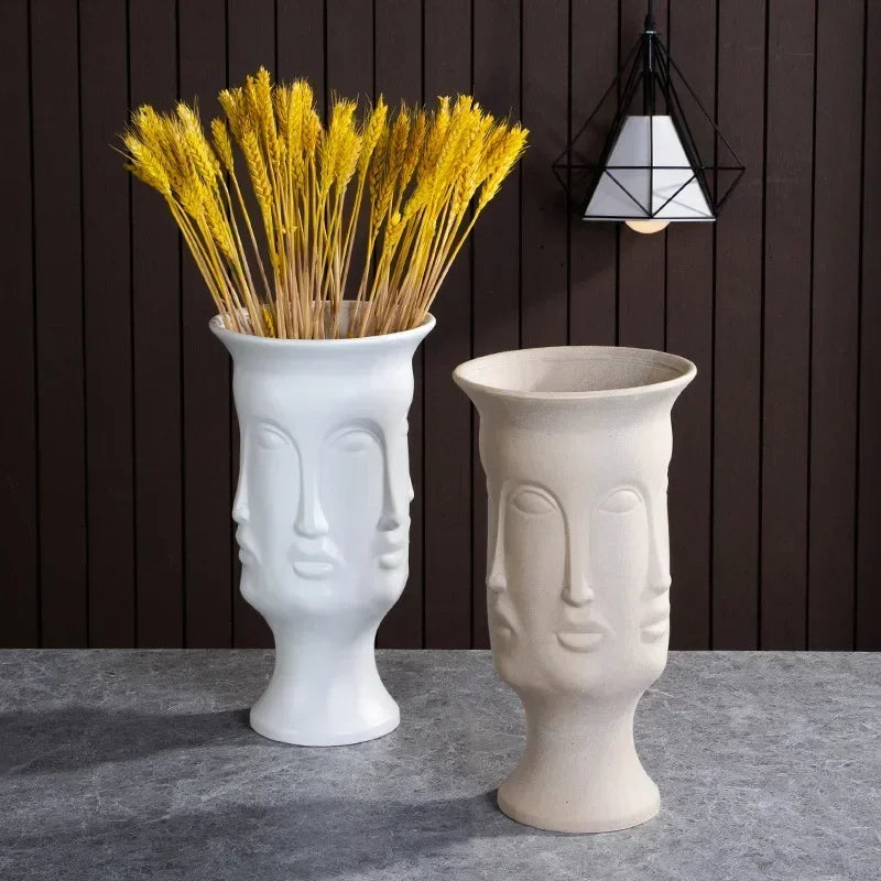 Tall goblet-style ceramic vases with elongated face designs in natural and white, styled with dried grasses
