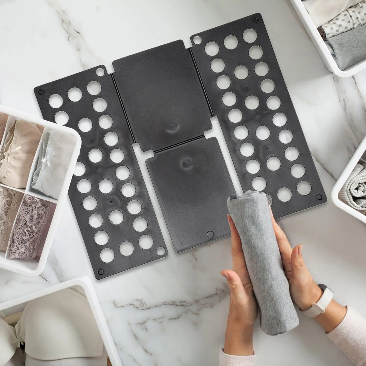 Hands folding a grey shirt near a black clothes folding board and divided storage bins