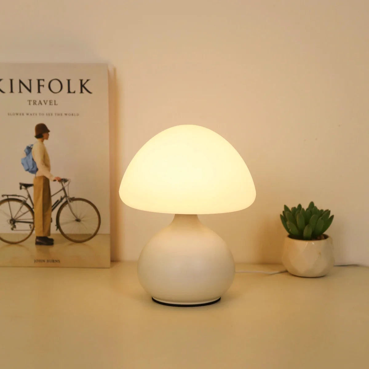 White mushroom table lamp glowing on a nightstand beside a book and succulent