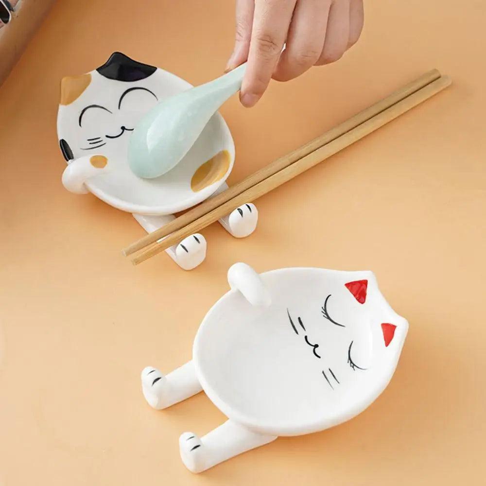 Hand setting a blue spoon onto a ceramic spoon rest with cat face design and yellow accents, next to another red-accent version
