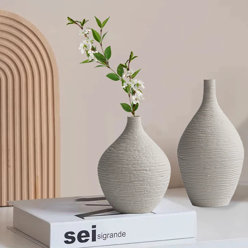 Pair of beige textured ceramic vases on a book, one holding faux white blossoms