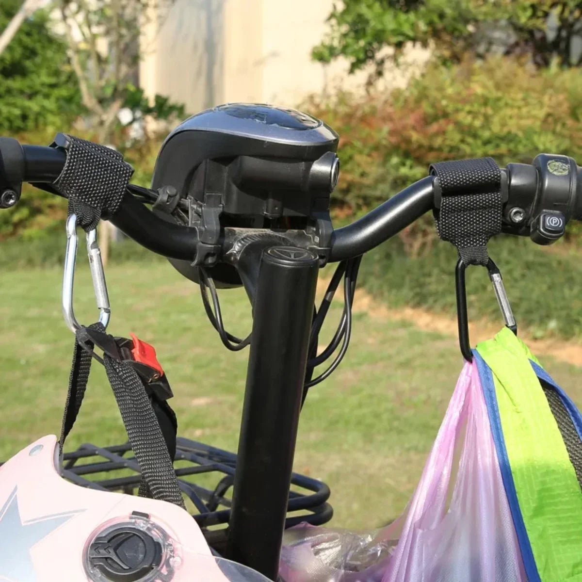 Two handlebar hooks installed on a black scooter handlebar, holding a helmet and shopping bags outdoors