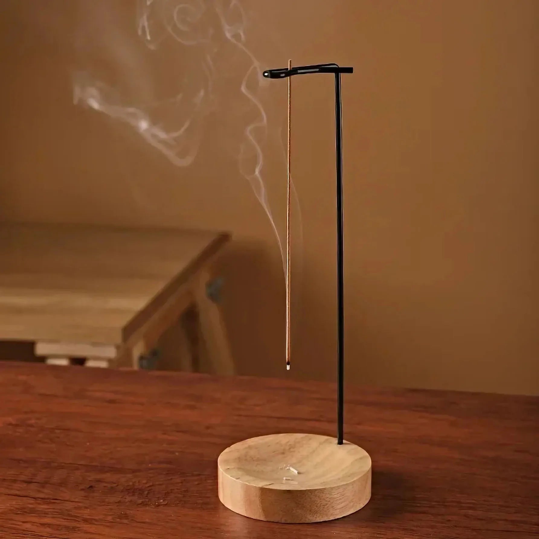 Wood incense stick holder with upright metal arm and round wood tray on a table, releasing incense smoke in a cozy room