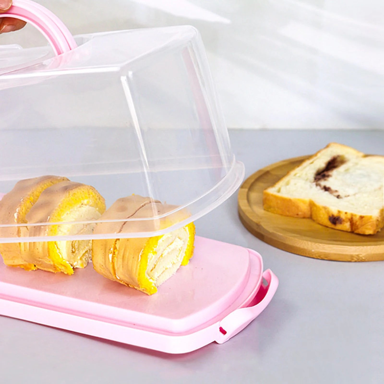 Plastic rectangular bread container with pink lid holding Swiss roll slices next to bread on a cutting board