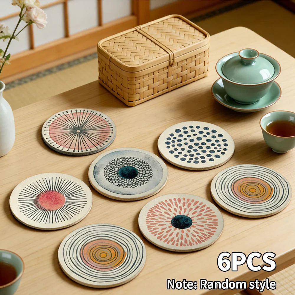 Six round art design coasters with abstract patterns on a wooden table beside tea cups and a woven storage box