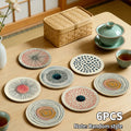 Six round art design coasters with abstract patterns on a wooden table beside tea cups and a woven storage box