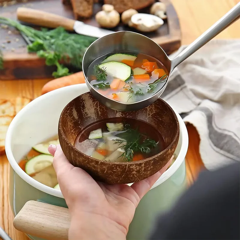 Hand holding coconut bowl being filled with vegetable soup in a kitchen setting