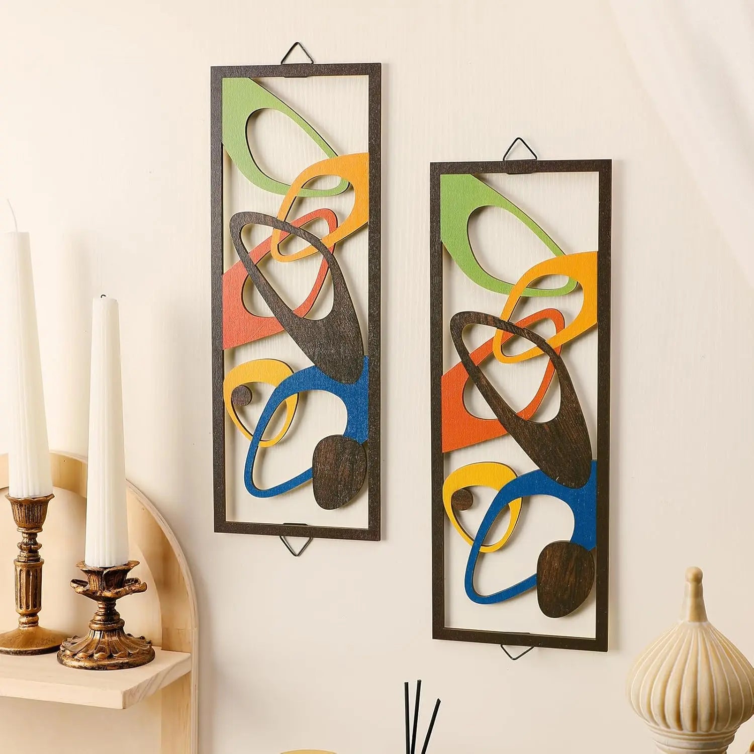 Pair of rectangular abstract wood wall art panels with colorful geometric shapes on a cream wall beside candlesticks