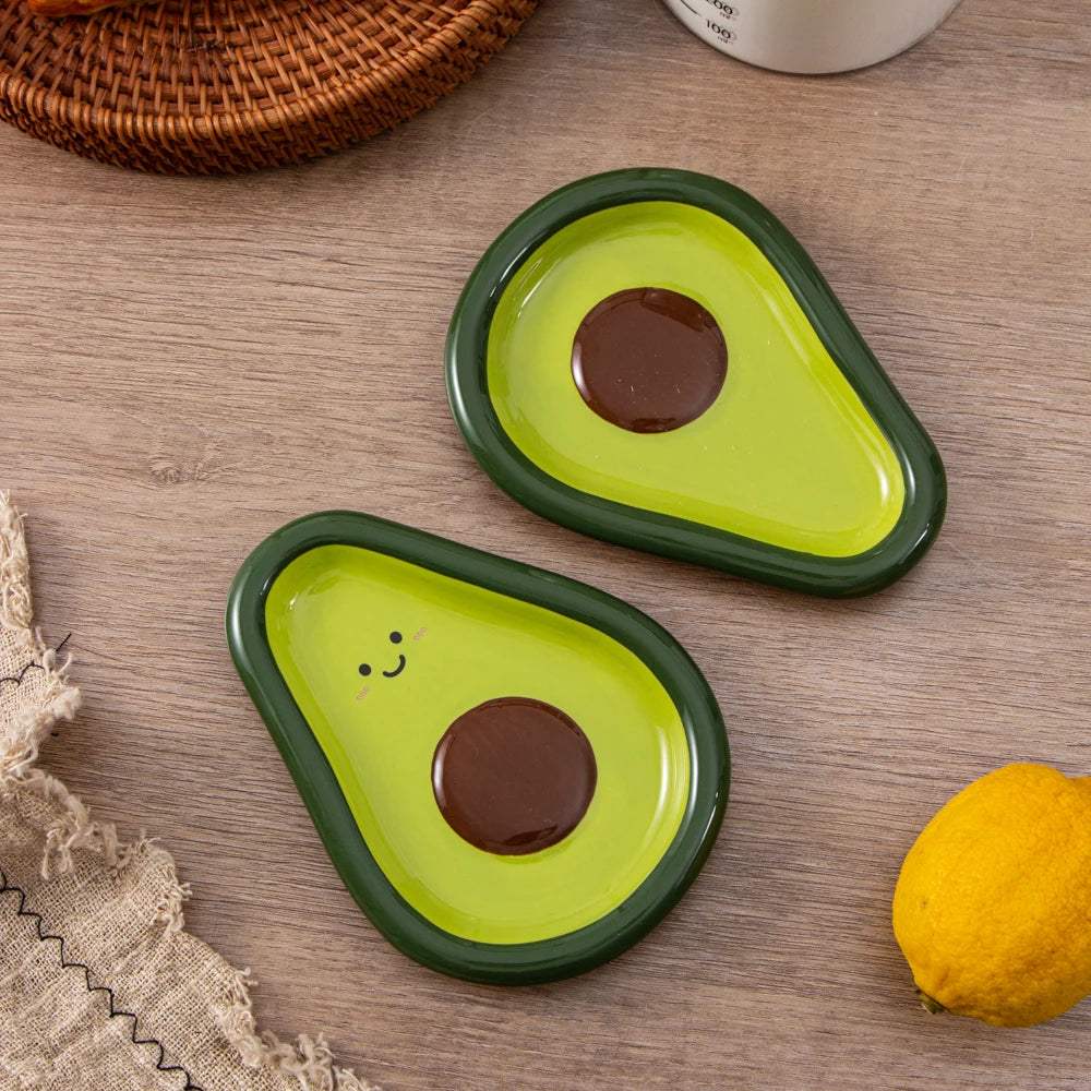 Two avocado-shaped ceramic spoon rests on a kitchen counter, one with a smiley face