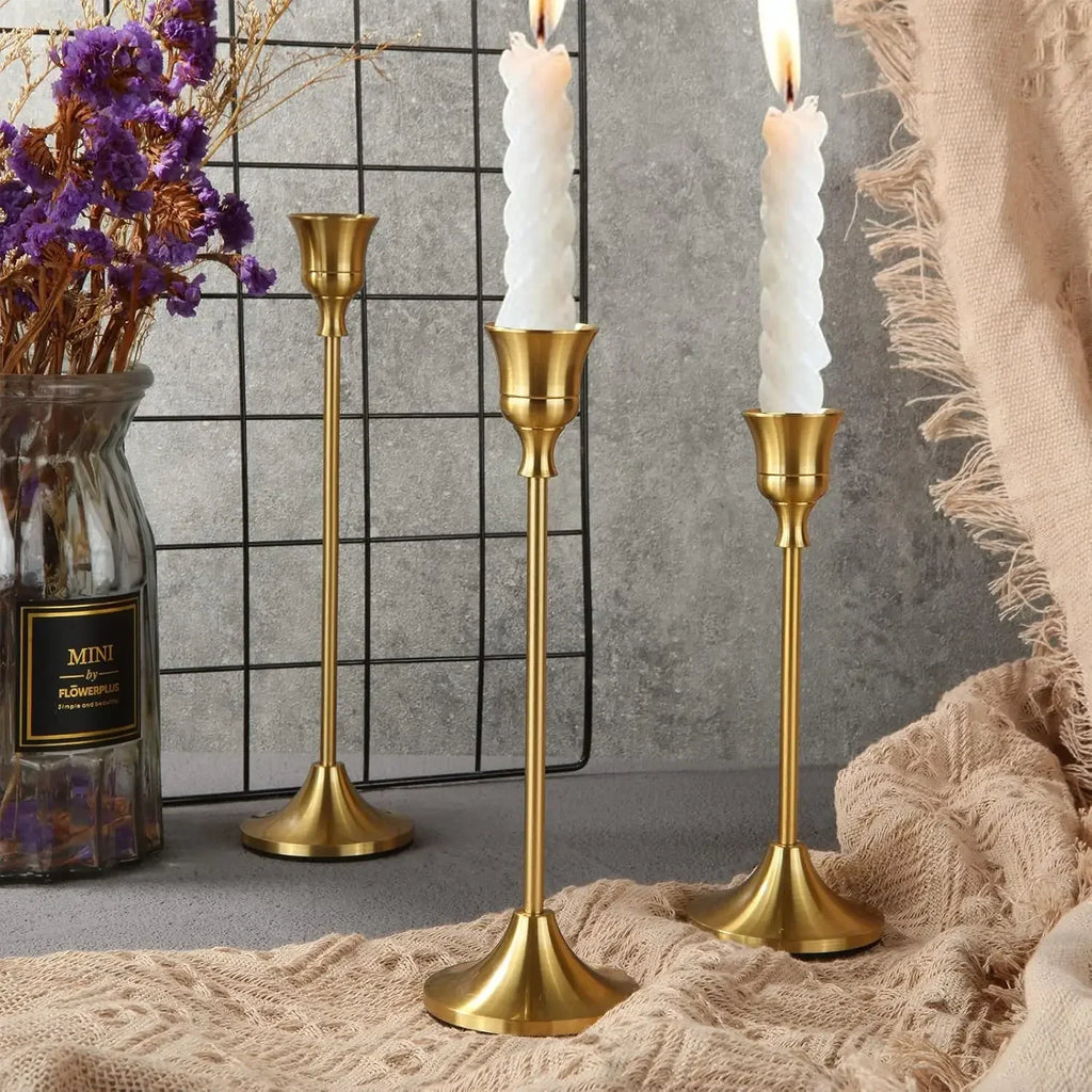 Three gold vintage-inspired metal taper candle holders on a tabletop, styled with dried flowers and cozy textiles.