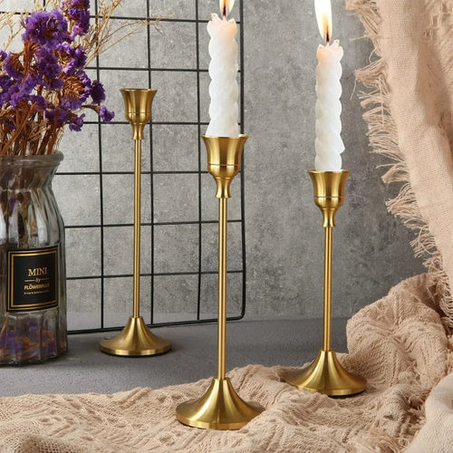 Three gold vintage-inspired metal taper candle holders on a tabletop, styled with dried flowers and cozy textiles.