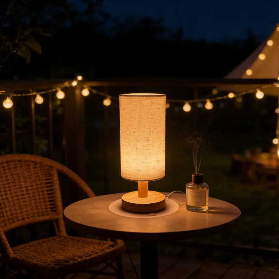 Wooden lamp with linen shade glowing on a small outdoor patio table, surrounded by string lights and a wicker chair at night