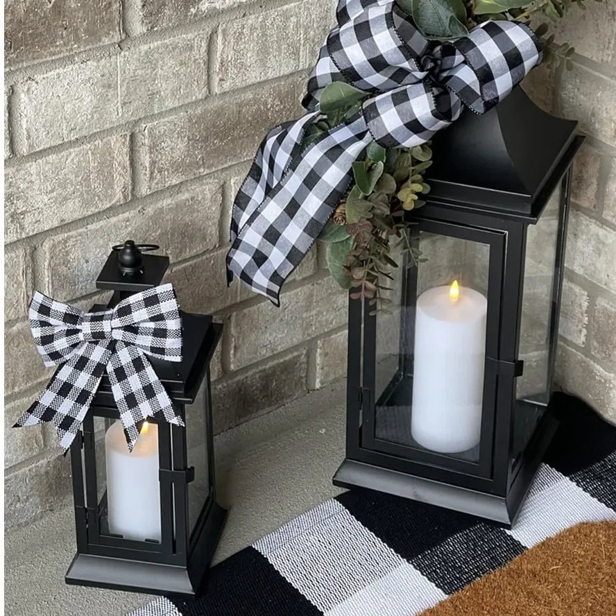 Two black lanterns with bows containing flameless LED pillar candles on a front porch with brick wall