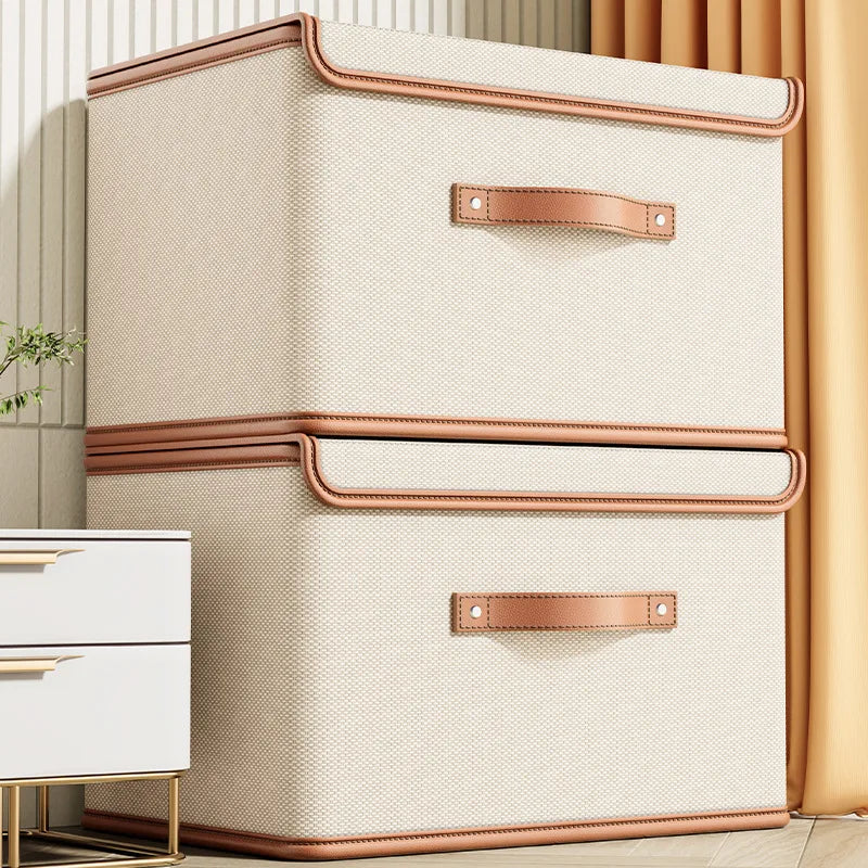 Two beige foldable lidded storage boxes with brown trim stacked in a bedroom corner