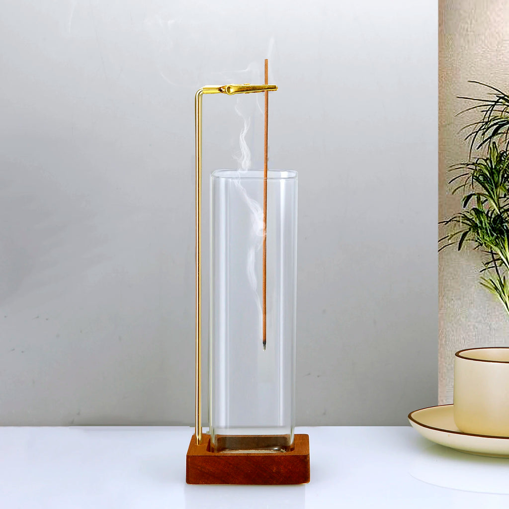 Glass and wood incense holder with brass clip holding a lit stick, displayed on a white surface beside a plant and teacup