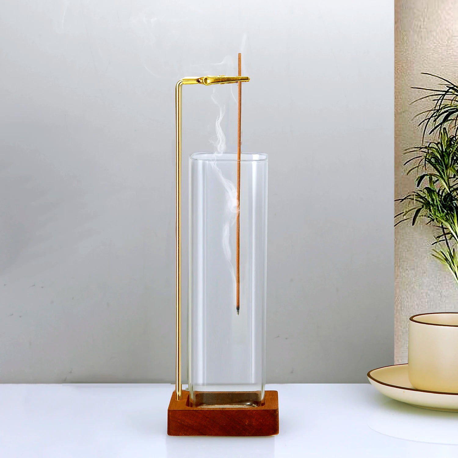 Glass and wood incense holder with brass clip holding a lit stick, displayed on a white surface beside a plant and teacup