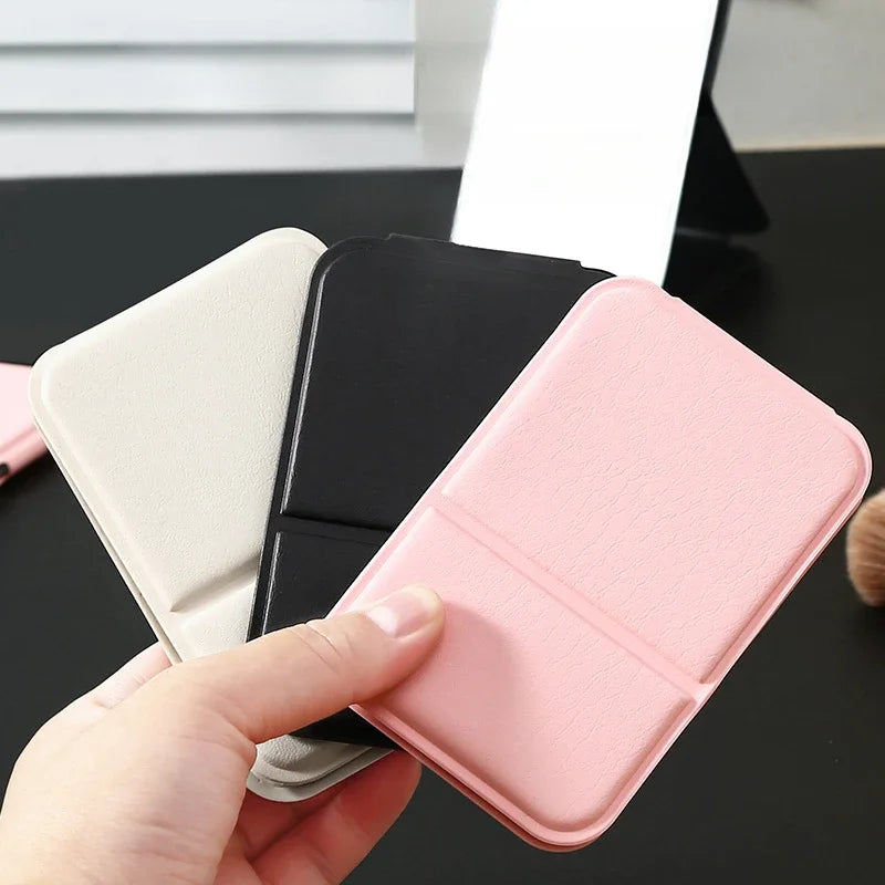 Hand holding three foldable mirrors in beige, black, and pink, with one mirror standing in the background