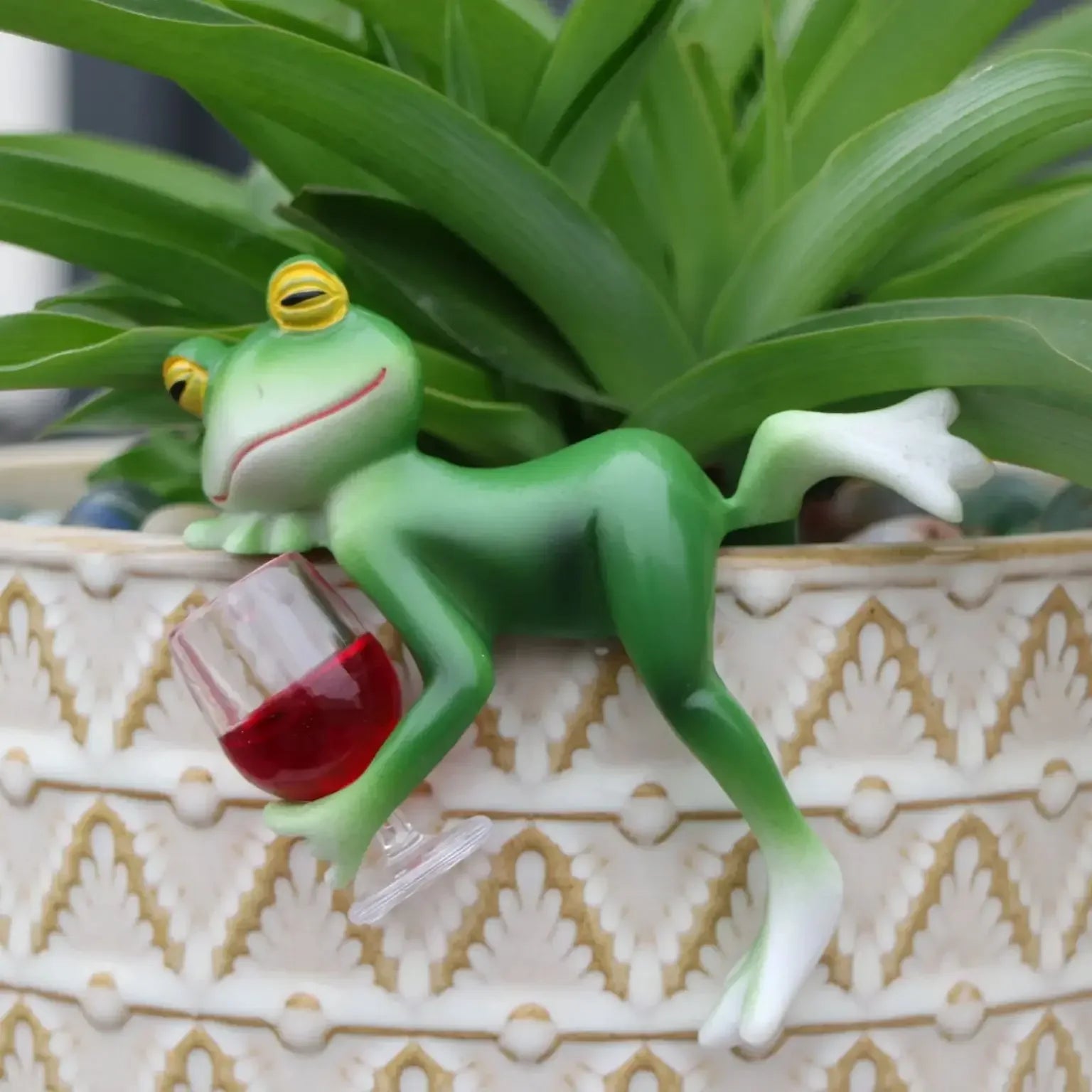 Reclining resin frog figure with wine glass displayed on an indoor planter with foliage