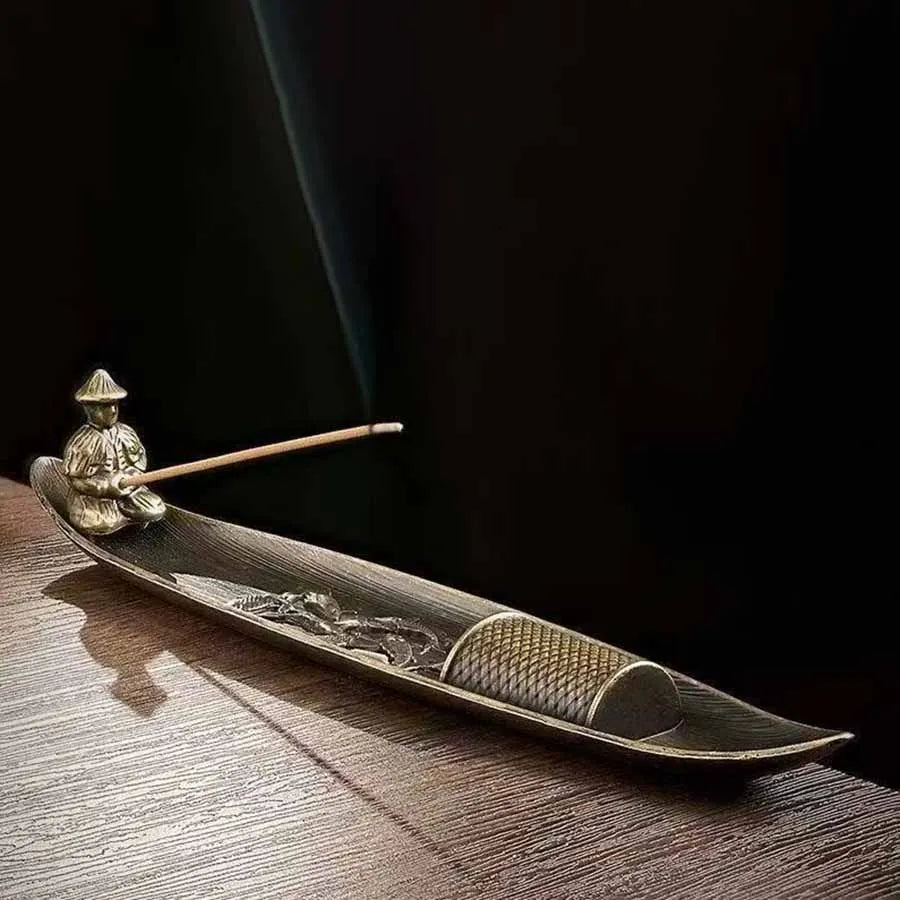 Decorative metal boat incense holder with seated figure at one end holding an incense stick