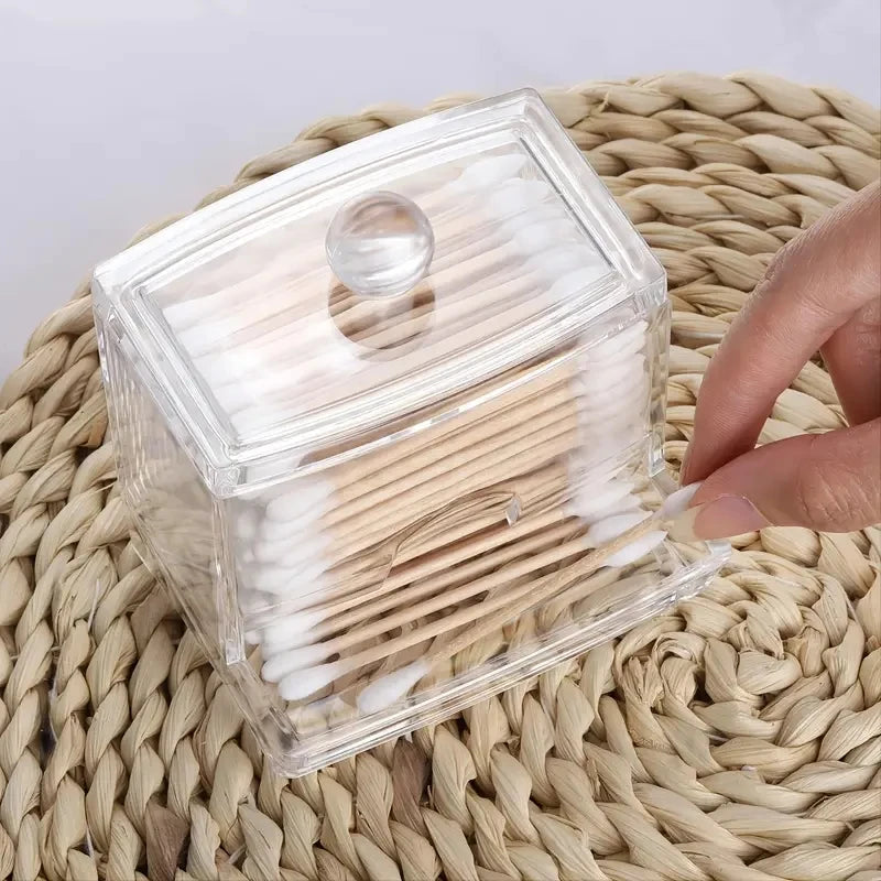 Clear lidded cotton swab holder with knob, sitting on a woven mat, hand reaching to grab a swab