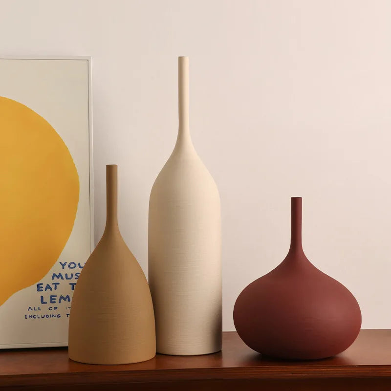 Mixed shapes of matte ceramic vases in beige, brown, and burgundy on a wood shelf