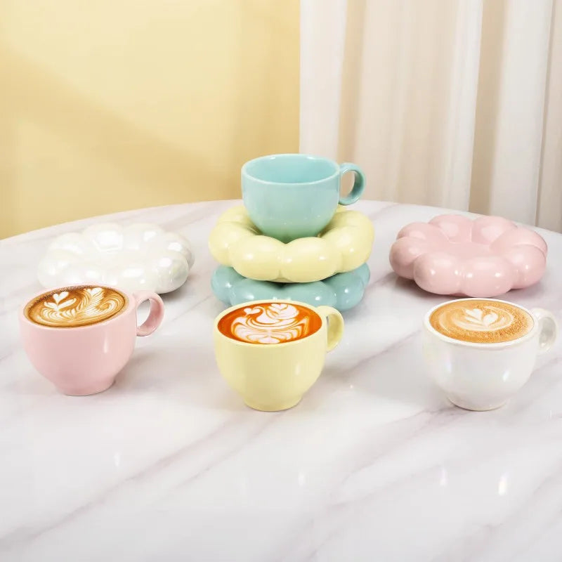 Pastel yellow, pink, blue, and white mugs with cloud saucers, some filled with latte art coffee drinks, stacked on a marble table