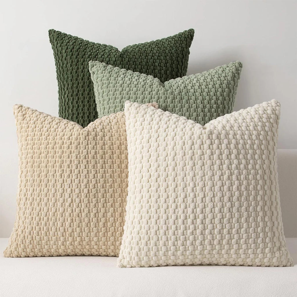A stack of four square textured corduroy pillow covers in beige and green tones on a light sofa, showing soft woven detail