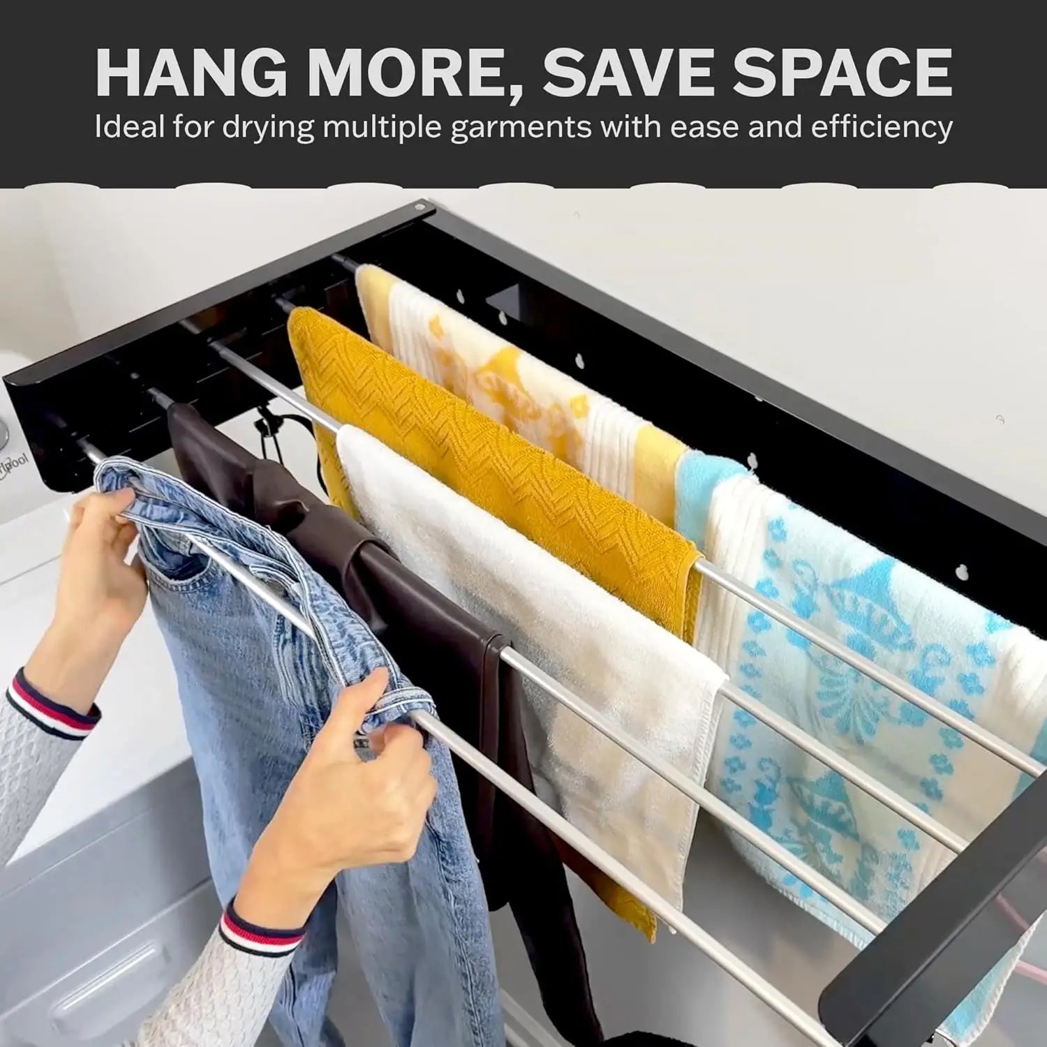 Person hanging jeans on a black wall-mounted drying rack with multiple towels and clothes above a washing machine