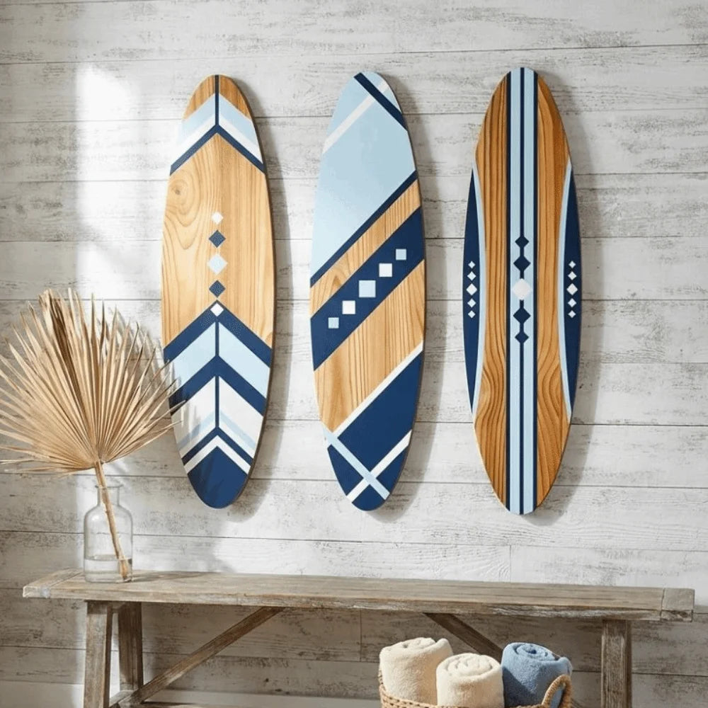 Three wooden surfboard wall art pieces with blue, white, and natural accent colors on a painted white wood wall with bench and rolled towels.