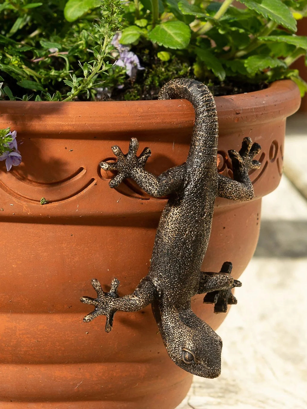 Resin gecko figurine hanging on the side of a terracotta flower pot outdoors