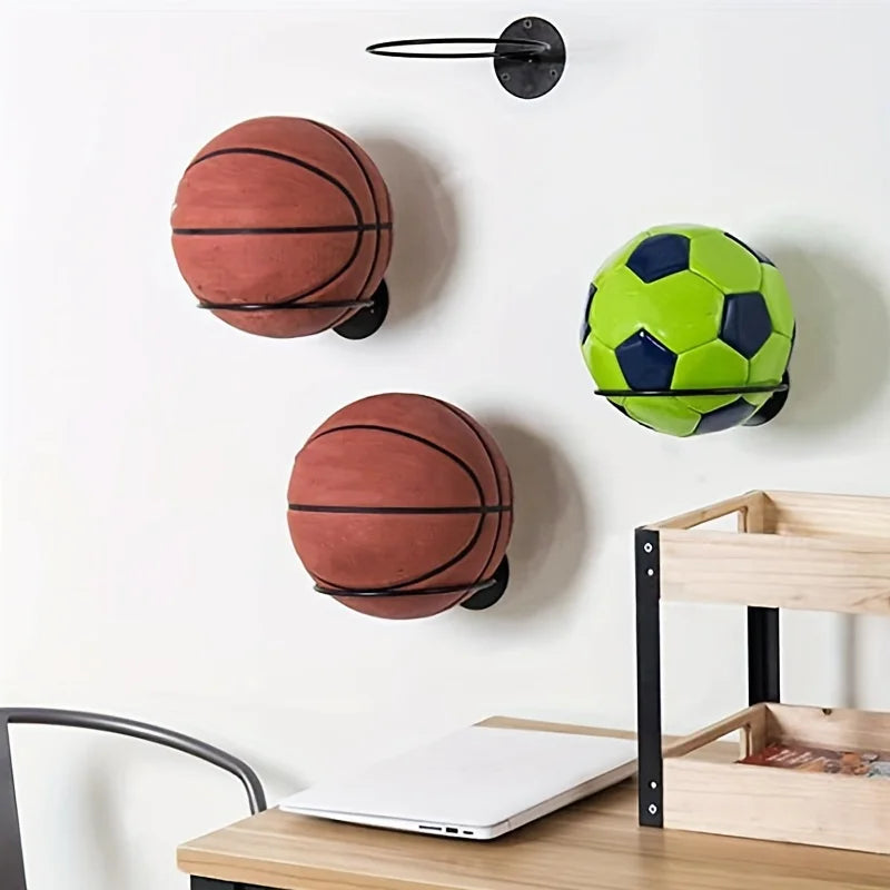 Three wall-mounted metal sports ball racks holding two basketballs and one soccer ball above a desk in a home setting