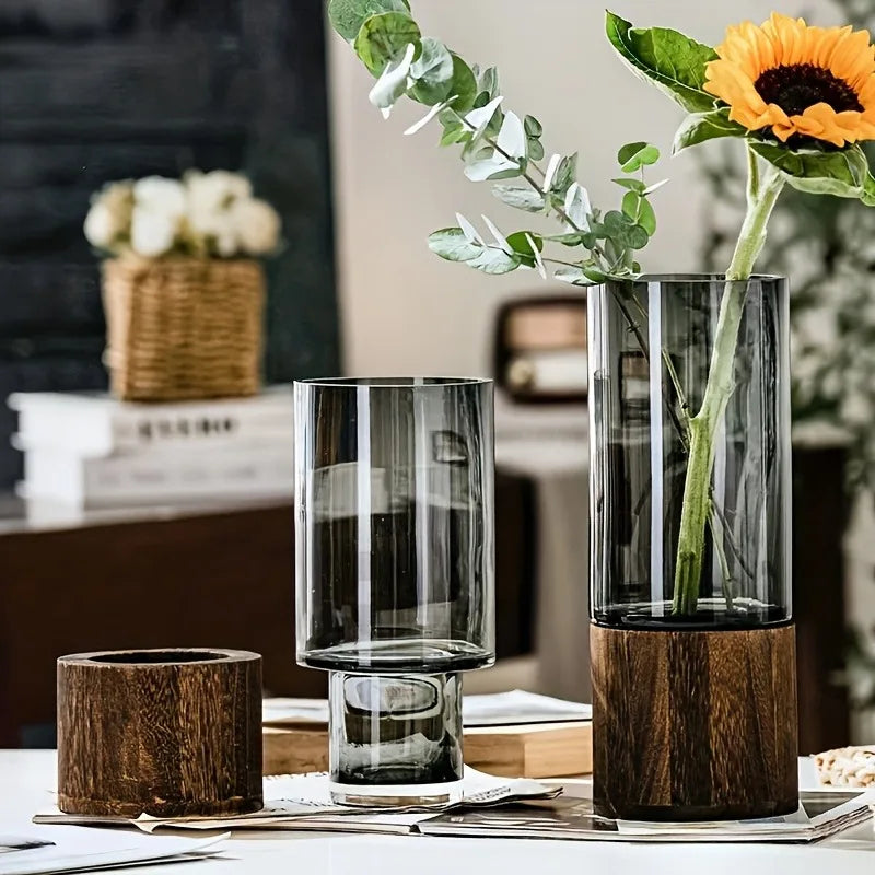 Two smoky glass vases with wooden bases, one holding greenery and a sunflower