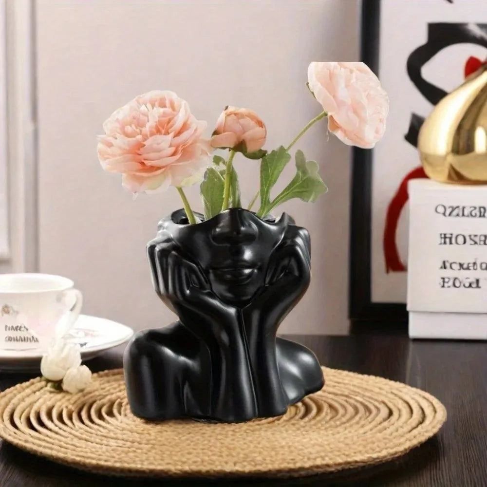 Black face sculpture vase with pink flowers on a woven placemat, styled on a desk