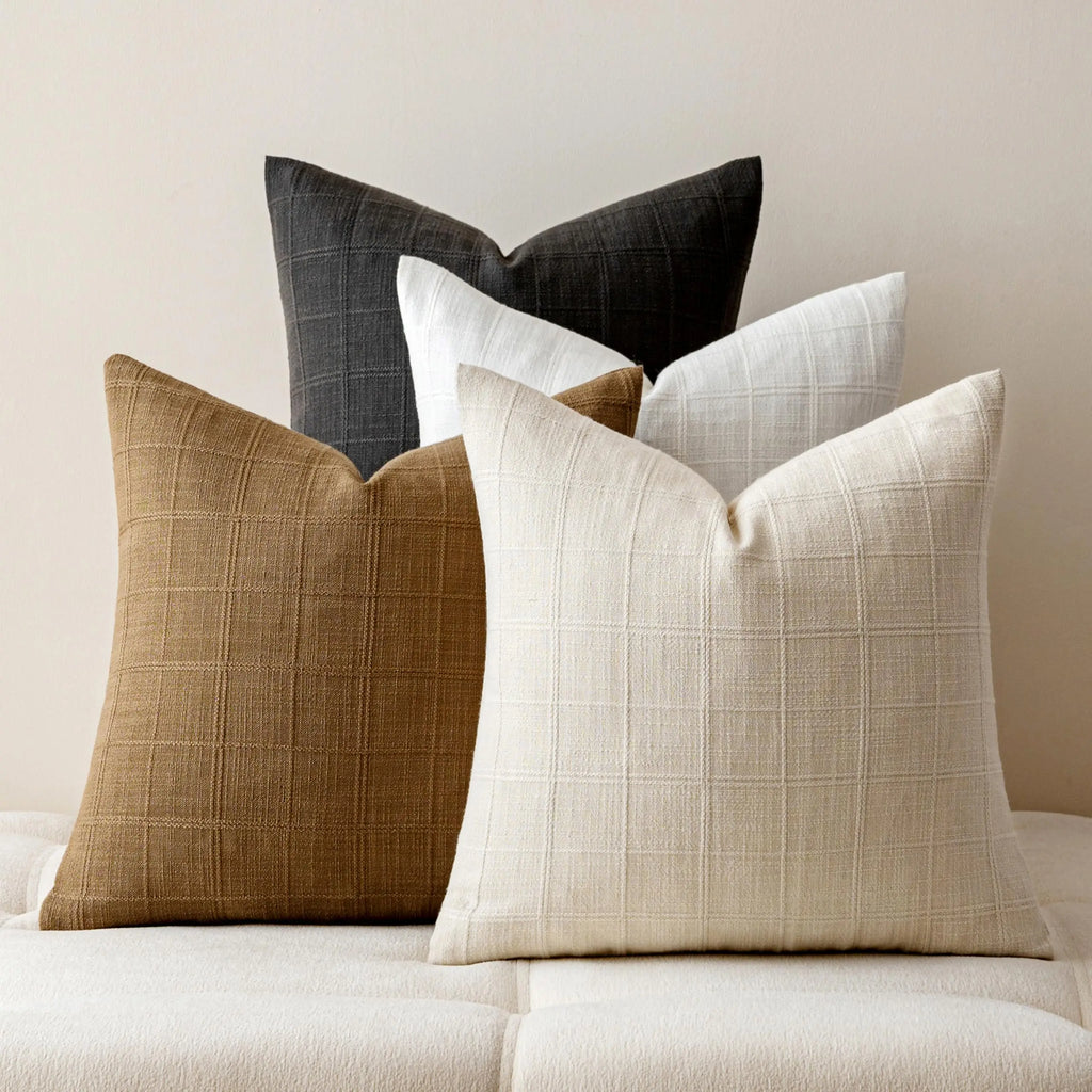 Stack of four linen plaid throw pillow covers in black, white, brown, and beige on a soft bench