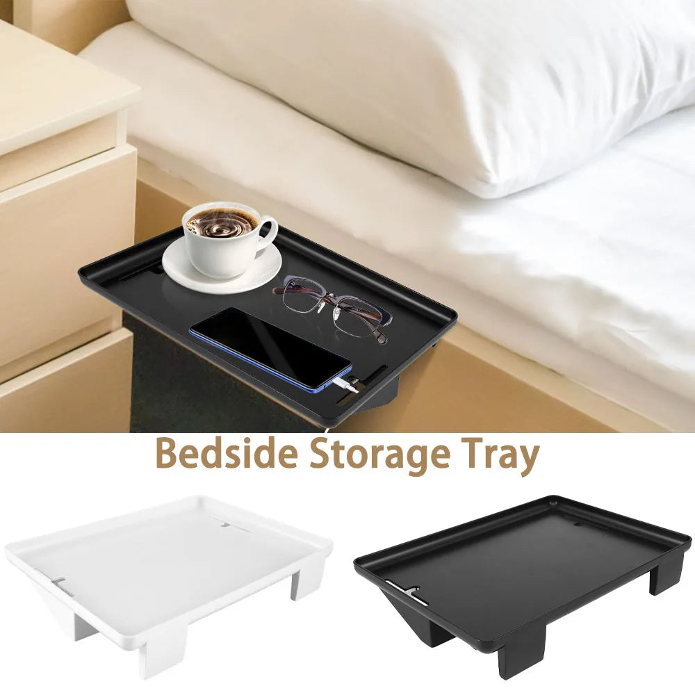 Black clip-on bedside storage tray attached to bed rail, holding coffee cup, glasses, and phone