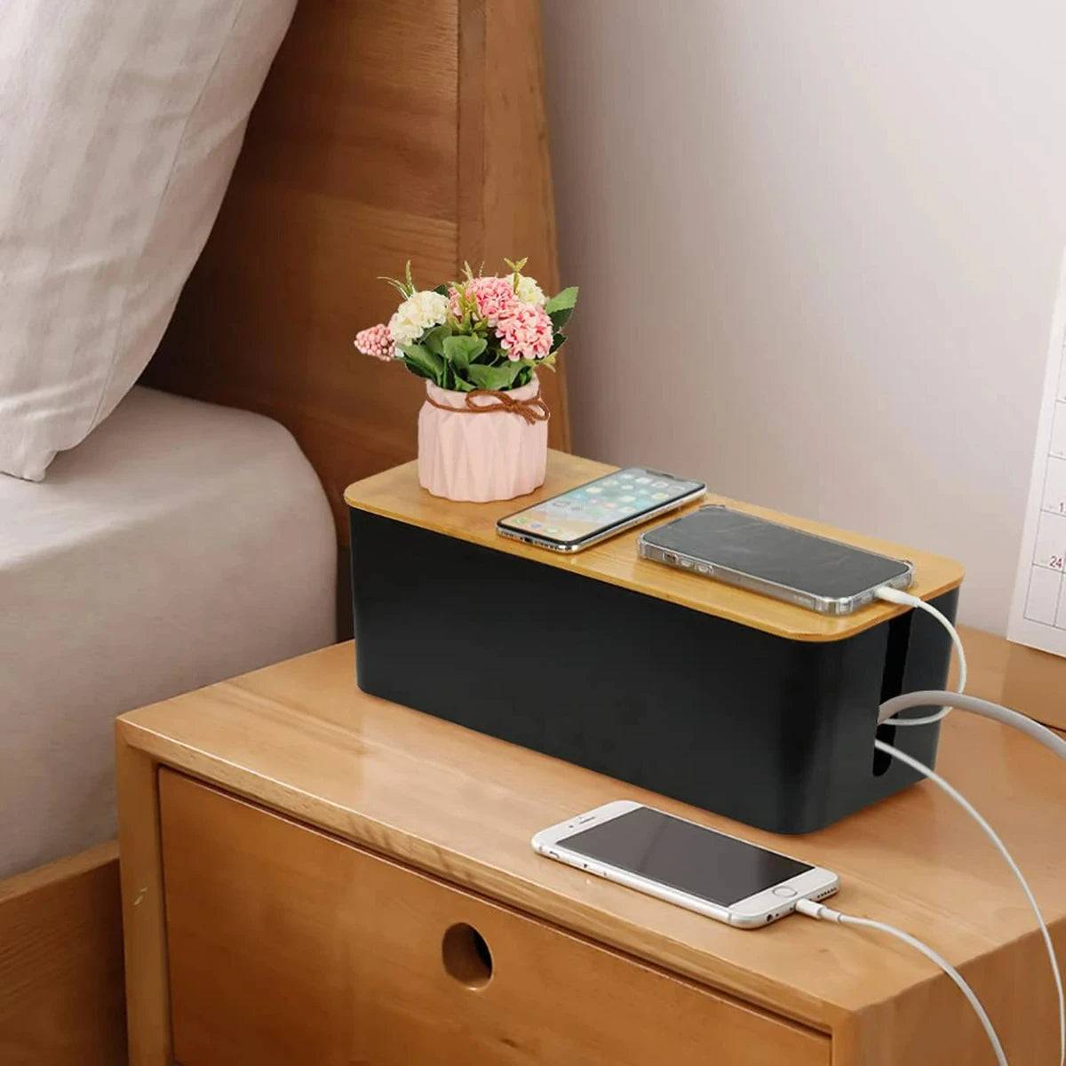 Black cable management box with bamboo lid on a nightstand, phones and flower pot on top