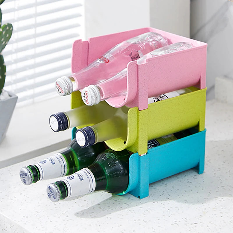 three stackable bottle organizer racks in pink, green, and blue, holding bottles on a kitchen countertop