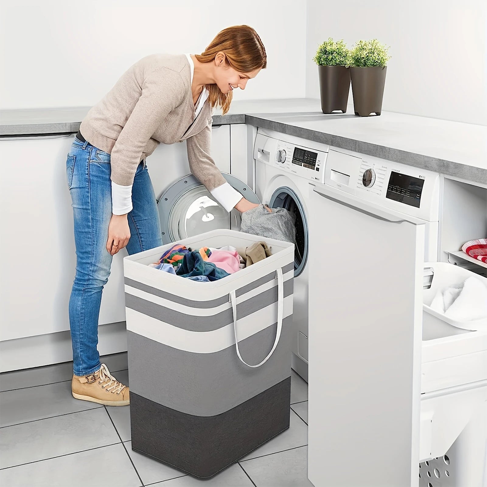 Person loading clothes from a striped rectangular laundry hamper into a front-load washing machine in a modern laundry room