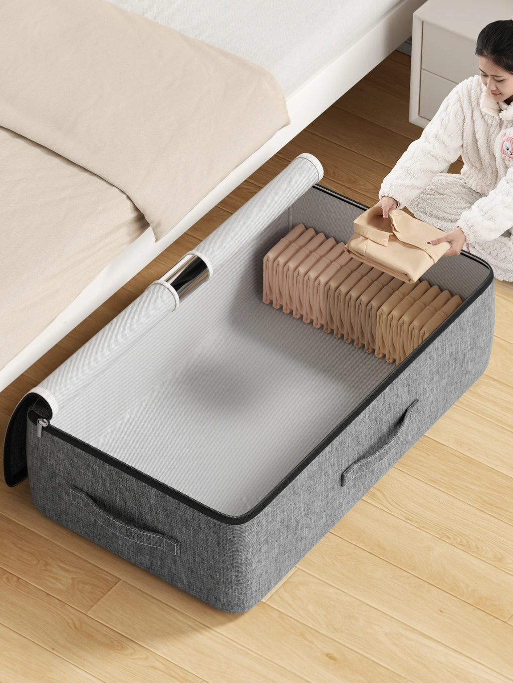Grey underbed storage bag open on bedroom floor, being filled with folded clothes