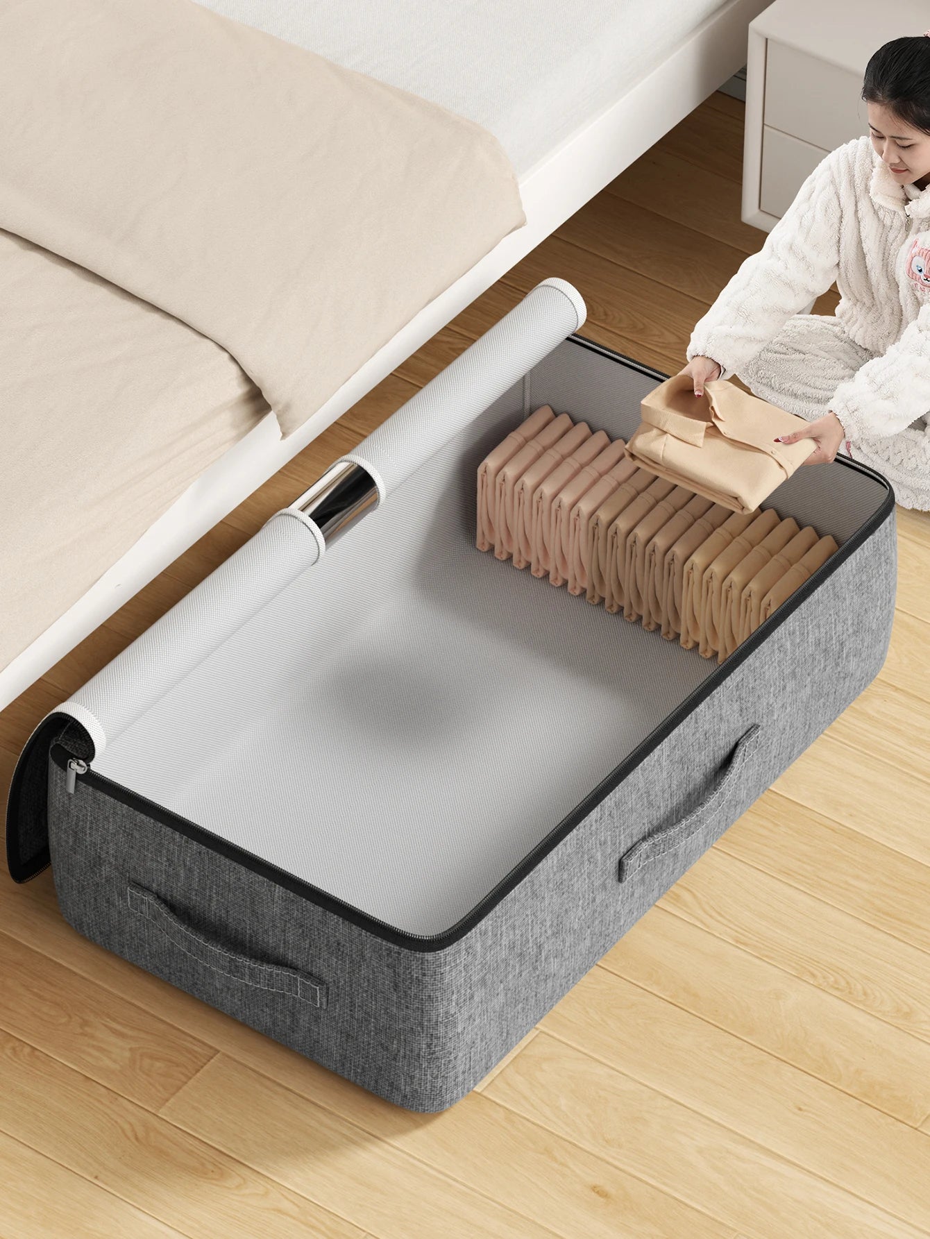 Grey underbed storage bag open on floor beside bed, person packing folded clothes inside